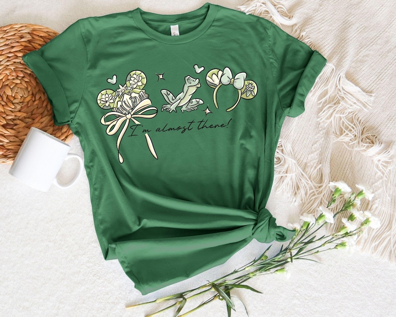 Puede incluir: Camiseta verde con un dise&ntilde;o caprichoso que presenta una rana, un lazo y elementos florales. El texto "I'm almost there!" est&aacute; impreso debajo del gr&aacute;fico. La camiseta est&aacute; anudada en la parte inferior.