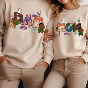 May include: Two beige sweatshirts with a graphic print of the words "DAD" and "MOM" in colourful letters. The letters are decorated with cartoon characters from the film Moana.