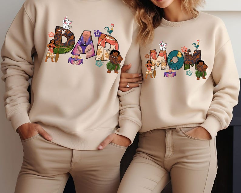 May include: Two beige sweatshirts with a graphic print of the words "DAD" and "MOM" in colourful letters. The letters are decorated with cartoon characters from the film Moana.
