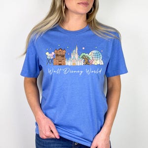 May include: A blue t-shirt featuring a colorful graphic of Walt Disney World landmarks, including a tower, castle, and Spaceship Earth. The words "Walt Disney World" are written in a script font below the graphic. The shirt is made of a soft material.
