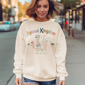 May include: Cream-colored sweatshirt with the text "Animal Kingdom" in rainbow colors. Below the text is a graphic of cartoon animals and the words "est. 1998 Orlando". The sweatshirt is being worn by a person.