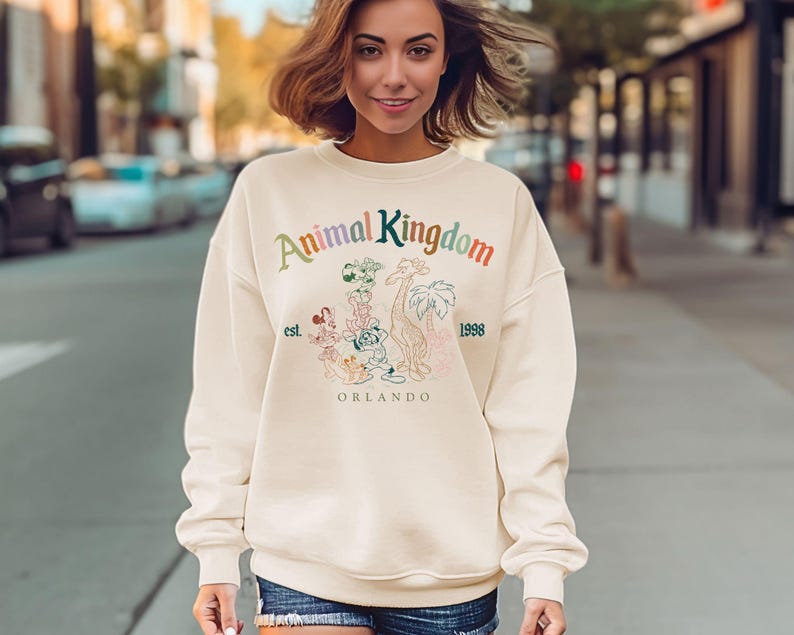 May include: Cream-colored sweatshirt with the text "Animal Kingdom" in rainbow colors. Below the text is a graphic of cartoon animals and the words "est. 1998 Orlando". The sweatshirt is being worn by a person.