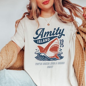 以下が含まれることがあります： クリーム色のTシャツ。サメのグラフィックと「Amity Island 1975」の文字がネイビーと赤でプリントされています。「You're gonna need a bigger boat!」のテキストがサメの下に印刷されています。