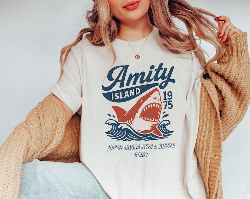 以下が含まれることがあります： クリーム色のTシャツ。サメのグラフィックと「Amity Island 1975」の文字がネイビーと赤でプリントされています。「You're gonna need a bigger boat!」のテキストがサメの下に印刷されています。