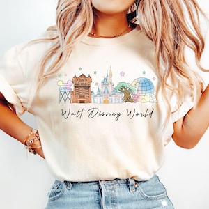 May include: Cream-colored t-shirt featuring a colorful illustration of Walt Disney World landmarks, including a water tower, castle, and Spaceship Earth. The words "Walt Disney World" are written in a script font below the graphic.