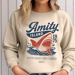 以下が含まれることがあります： ベージュ色のスウェットシャツ。サメのグラフィックと「Amity Island 1975」の文字が紺色と赤色で描かれています。サメの下には「You're gonna need a bigger boat！」の文字。デザインには海の波が含まれています。