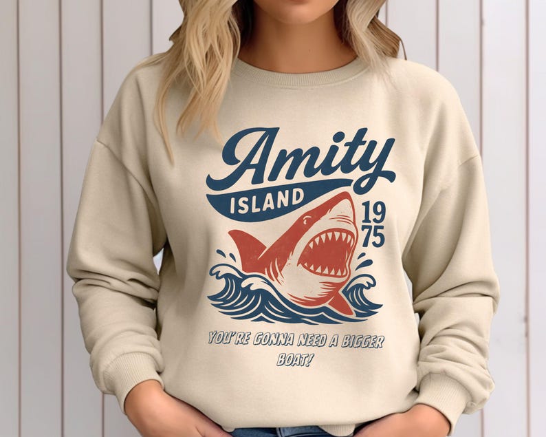 以下が含まれることがあります： ベージュ色のスウェットシャツ。サメのグラフィックと「Amity Island 1975」の文字が紺色と赤色で描かれています。サメの下には「You're gonna need a bigger boat！」の文字。デザインには海の波が含まれています。