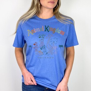 May include: A blue t-shirt with the words "Animal Kingdom" in colorful letters. The shirt features a graphic of cartoon characters and the text "est. 1998 Orlando". The shirt is made of a soft material.