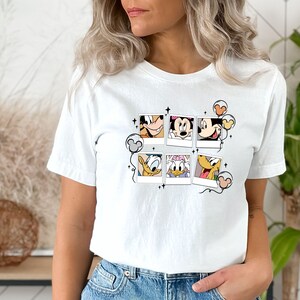 May include: White t-shirt with a graphic of Mickey Mouse, Minnie Mouse, Donald Duck, and Goofy in Polaroid frames with balloons. The graphic is in black and white with pops of color in the balloons.