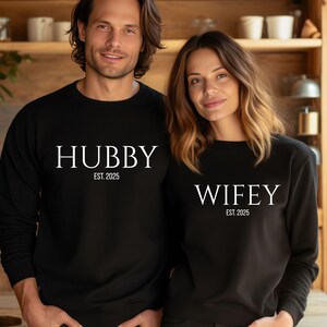 May include: Matching black sweatshirts with white text that reads "HUBBY EST. 2025" and "WIFEY EST. 2025". The sweatshirts are long-sleeved and worn by a couple. The background is a wooden kitchen.