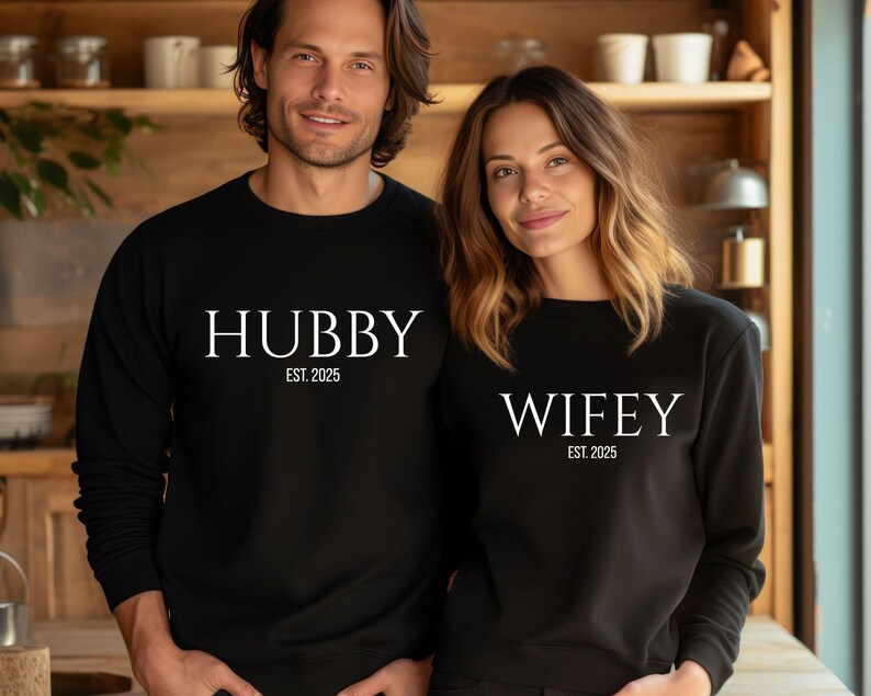 May include: Matching black sweatshirts with white text that reads "HUBBY EST. 2025" and "WIFEY EST. 2025". The sweatshirts are long-sleeved and worn by a couple. The background is a wooden kitchen.