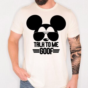 May include: A cream-colored short-sleeve t-shirt featuring a black graphic of a mouse silhouette wearing sunglasses. Below the graphic, the text reads TALK TO ME GOOF in bold black capital letters, flanked by two horizontal lines on each side.
