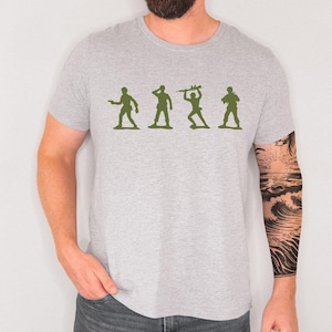 May include: A light grey t-shirt with a green silhouette of four toy soldiers in different poses. The shirt is made of a soft material and has a crew neck. The person wearing the shirt has a tattoo on their arm.