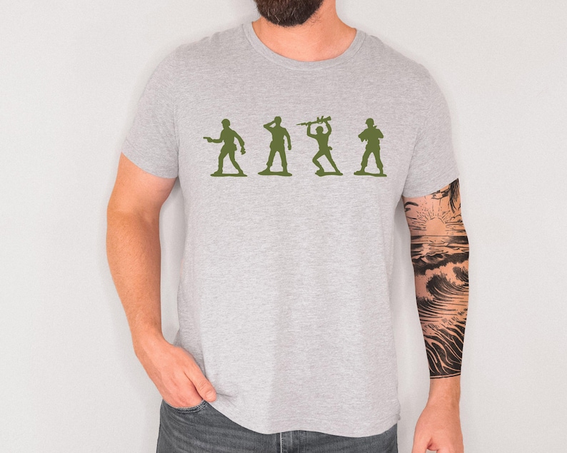 May include: A light grey t-shirt with a green silhouette of four toy soldiers in different poses. The shirt is made of a soft material and has a crew neck. The person wearing the shirt has a tattoo on their arm.