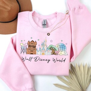 May include: A light pink sweatshirt featuring a colorful graphic of Walt Disney World landmarks, including Cinderella's castle and Spaceship Earth. The words "Walt Disney World" are written in a script font below the graphic. A gold necklace is draped on the sweatshirt.