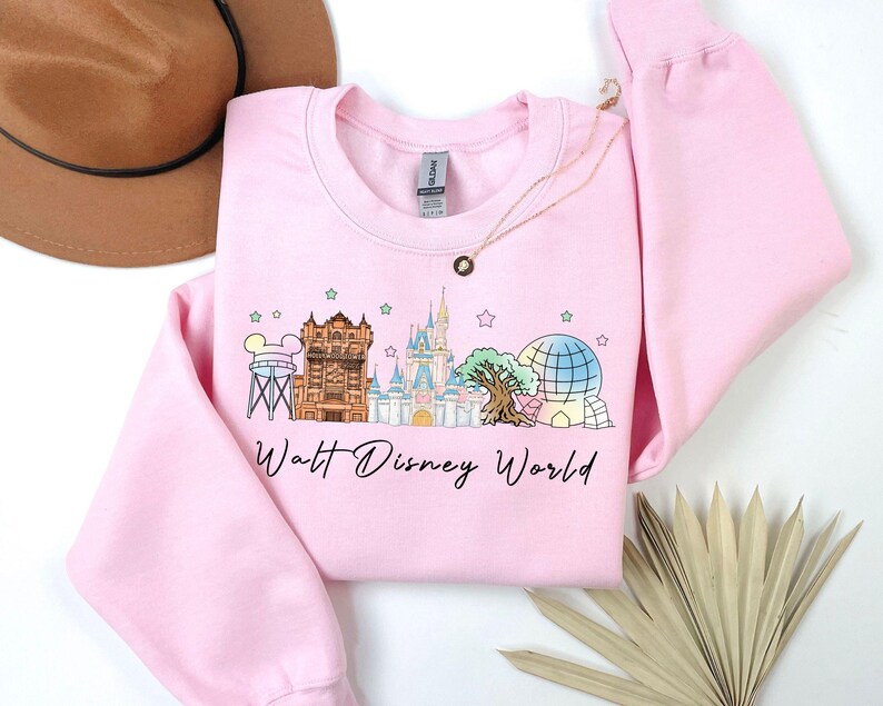 May include: A light pink sweatshirt featuring a colorful graphic of Walt Disney World landmarks, including Cinderella's castle and Spaceship Earth. The words "Walt Disney World" are written in a script font below the graphic. A gold necklace is draped on the sweatshirt.