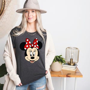 Puede incluir: Una camiseta gris oscura con una imagen de dibujos animados de Minnie Mouse con un lazo rojo con lunares blancos.