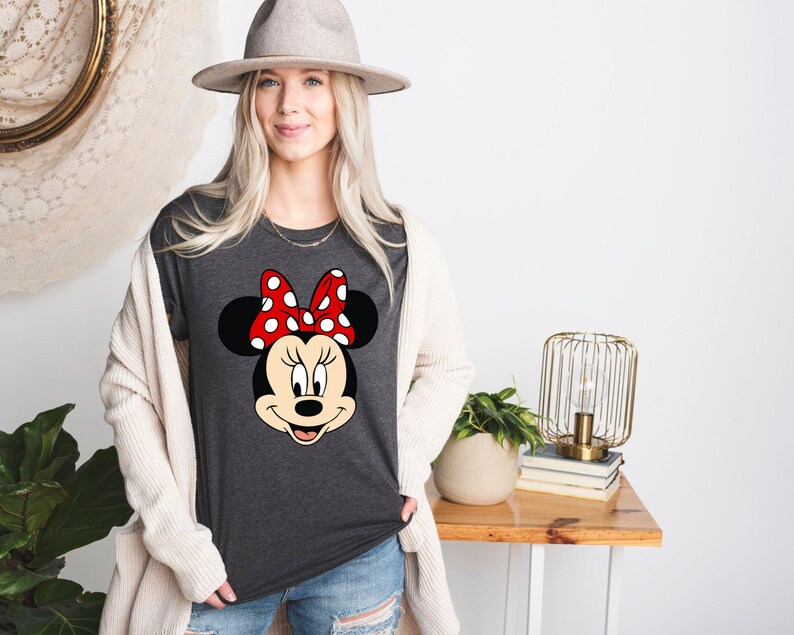 Puede incluir: Una camiseta gris oscura con una imagen de dibujos animados de Minnie Mouse con un lazo rojo con lunares blancos.