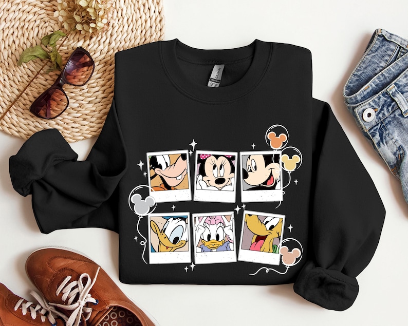 May include: Black sweatshirt with a graphic print of Mickey Mouse, Minnie Mouse, Donald Duck, Goofy, and Pluto in Polaroid frames with balloons. The characters are smiling and looking at the camera.