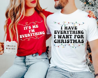 May include: Two t-shirts, one red and one white, with Christmas-themed text. The red t-shirt says "IT'S ME I'M EVERYTHING", and the white one says "I HAVE EVERYTHING I WANT FOR CHRISTMAS". Both have a string of lights.