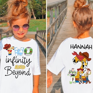 Puede incluir: Camiseta blanca con el texto "Two infinity and Beyond" y un gráfico de dibujos animados. La parte posterior de la camiseta tiene el nombre "Hannah" y un gráfico de "Toy Story". La camiseta la lleva una niña pequeña con gafas de sol.