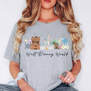 May include: A light gray t-shirt featuring a colorful illustration of the Walt Disney World skyline. The design includes iconic landmarks like Cinderella's Castle and Spaceship Earth, with the words "Walt Disney World" in a script font.