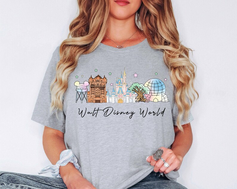 May include: A light gray t-shirt featuring a colorful illustration of the Walt Disney World skyline. The design includes iconic landmarks like Cinderella's Castle and Spaceship Earth, with the words "Walt Disney World" in a script font.