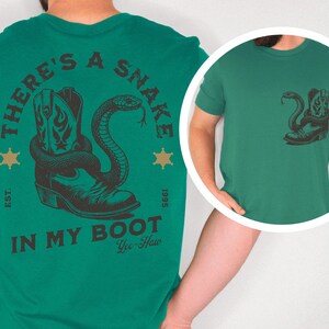 Puede incluir: Camiseta verde con un dise&ntilde;o de una bota de vaquero y una serpiente. El texto "THERE'S A SNAKE IN MY BOOT" est&aacute; impreso encima y debajo del dise&ntilde;o. La camiseta tambi&eacute;n tiene el texto "Yee-Haw" y "EST. 1995".