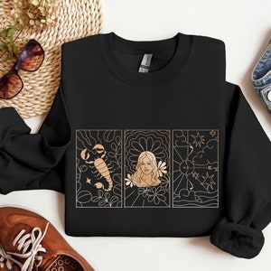 Pode incluir: Camisola preta de gola redonda com um gráfico dourado e bege de três painéis. Os painéis retratam um escorpião, o rosto de uma mulher e um arco e flecha. A camisola é feita de um material macio.
