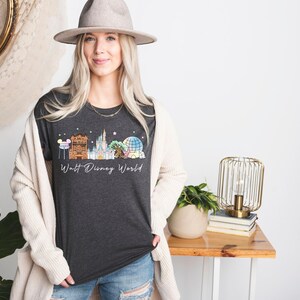 May include: Dark gray t-shirt featuring a colorful illustration of the Walt Disney World skyline. The design includes iconic landmarks like Cinderella's Castle and Spaceship Earth, with the words "Walt Disney World" in a script font.