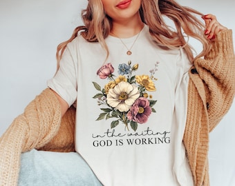 May include: Cream-colored t-shirt featuring a floral bouquet in shades of pink, yellow, purple, and blue. The text "in the waiting GOD IS WORKING" is printed below the flowers in a script font. The shirt is worn with a tan cardigan.