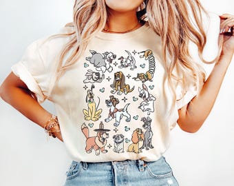 May include: Cream-colored t-shirt featuring a collection of cartoon dog illustrations in various poses. The dogs are in different colors and breeds, with some wearing hats or accessories. The design includes small heart and star accents.