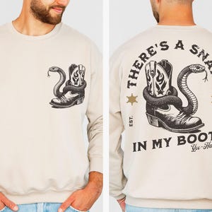 Puede incluir: Sudadera beige con un gr&aacute;fico en blanco y negro de una bota de vaquero y una serpiente. La parte trasera de la sudadera muestra el texto "THERE'S A SNAKE IN MY BOOT". Cuello redondo y mangas largas.