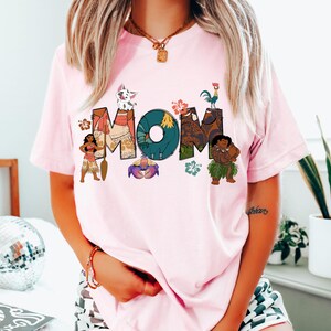May include: Pink t-shirt featuring a graphic of the word "MOM" made up of characters from the animated film Moana. The characters include Maui, Pua, Hei Hei, Moana, and a crab.
