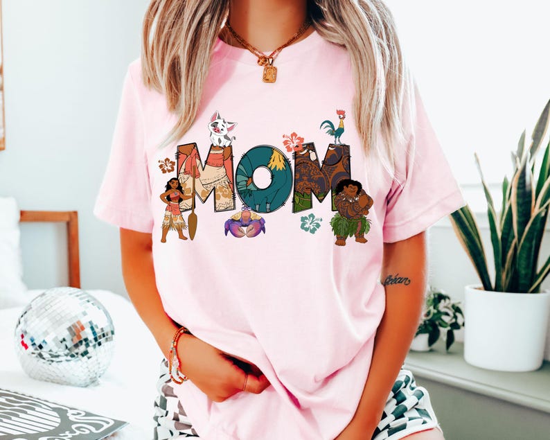May include: Pink t-shirt featuring a graphic of the word "MOM" made up of characters from the animated film Moana. The characters include Maui, Pua, Hei Hei, Moana, and a crab.