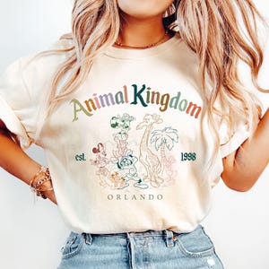 May include: Cream-colored t-shirt featuring the text "Animal Kingdom" in a rainbow arch, with the words "est. 1998" below. The shirt has a graphic of cartoon animals, including a giraffe and palm trees. The word "ORLANDO" is printed at the bottom.