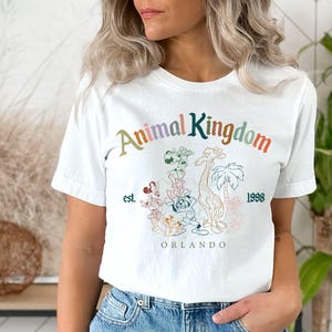 May include: White t-shirt featuring the text "Animal Kingdom" in a colorful, arched design. Below the text is a graphic of a giraffe, palm trees, and other animals. The shirt also includes the text "est. 1998" and "ORLANDO".