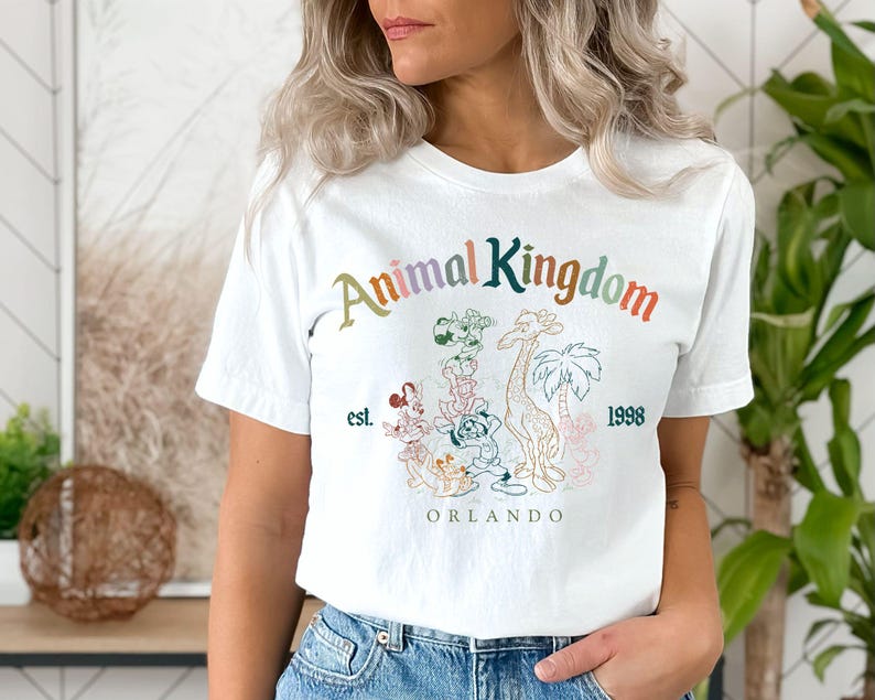 May include: White t-shirt featuring the text "Animal Kingdom" in a colorful, arched design. Below the text is a graphic of a giraffe, palm trees, and other animals. The shirt also includes the text "est. 1998" and "ORLANDO".
