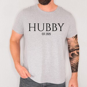 May include: A light gray t-shirt with the word "HUBBY" printed in black, with "EST. 2025" below it. The shirt is short-sleeved and made of a soft material. The person wearing the shirt has a tattoo on their arm.