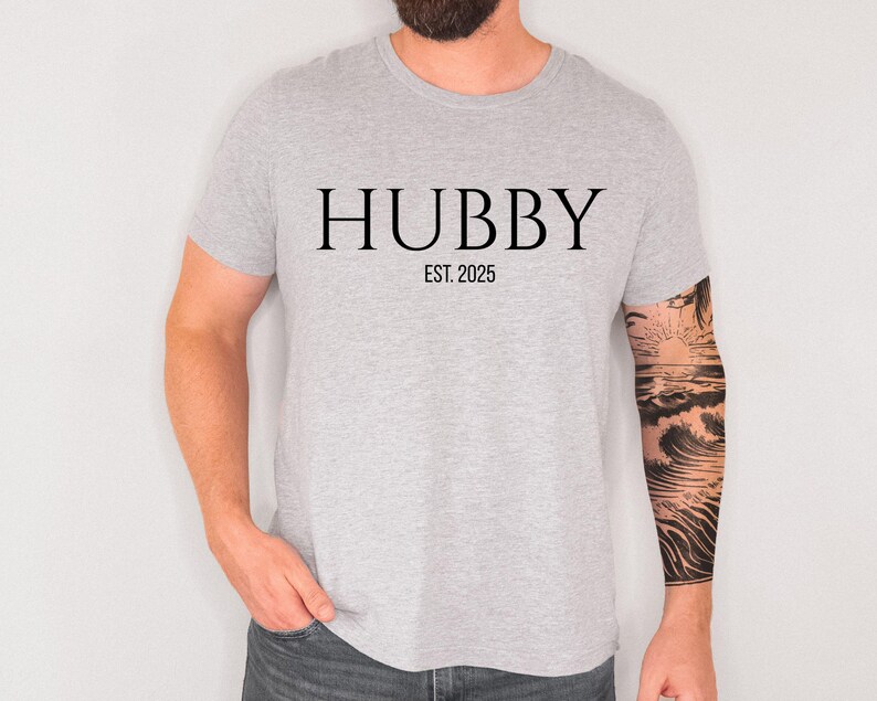 May include: A light gray t-shirt with the word "HUBBY" printed in black, with "EST. 2025" below it. The shirt is short-sleeved and made of a soft material. The person wearing the shirt has a tattoo on their arm.