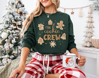 May include: A dark green sweatshirt with the text "The Cookie Crew" and gingerbread-themed graphics. The person is wearing red and white plaid pajama pants and holding a white mug with a Santa Claus design.