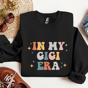 May include: Black sweatshirt with the text "In My Gigi Era" in colorful retro letters. The letters are outlined in white and have a glittery effect.