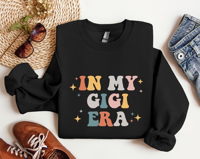 Puede incluir: Sudadera negra con el texto "In My Gigi Era" en letras retro de colores. Las letras est&aacute;n delineadas en blanco y tienen un efecto brillante.