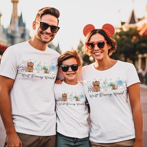 May include: Three people wearing white t-shirts with a colorful graphic of a cityscape and the text "Walt Disney World". The shirts have a crew neck and short sleeves. The people are smiling and wearing sunglasses.