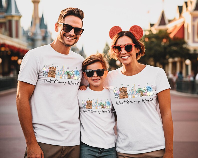 May include: Three people wearing white t-shirts with a colorful graphic of a cityscape and the text "Walt Disney World". The shirts have a crew neck and short sleeves. The people are smiling and wearing sunglasses.