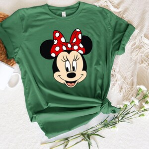 Puede incluir: Una camiseta verde con una imagen de dibujos animados de Minnie Mouse con un lazo rojo con lunares blancos.