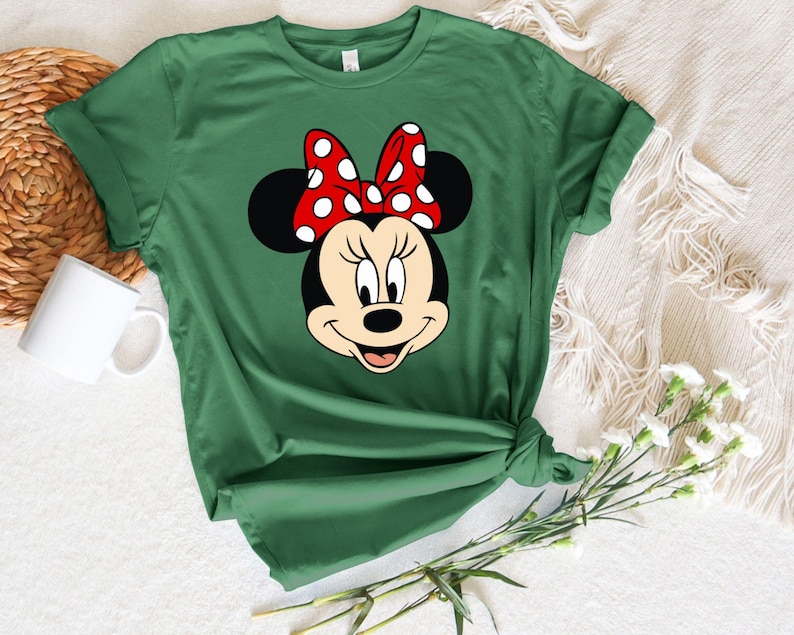 Puede incluir: Una camiseta verde con una imagen de dibujos animados de Minnie Mouse con un lazo rojo con lunares blancos.