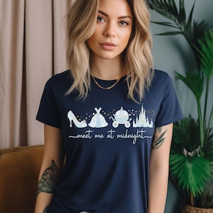 以下が含まれることがあります： ネイビーブルーのTシャツ。白いグラフィックはハイヒール、ドレス、馬車、お城。テキスト「meet me at midnight」は筆記体で書かれています。Tシャツはクルーネックです。