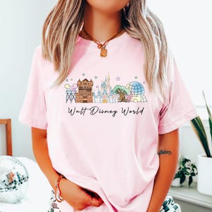 May include: Light pink t-shirt featuring a colorful illustration of Walt Disney World landmarks. The design includes the Tower of Terror, Cinderella Castle, Spaceship Earth, and the Tree of Life. The words "Walt Disney World" are written in a script font below the illustration.