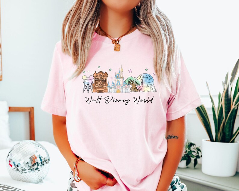 May include: Light pink t-shirt featuring a colorful illustration of Walt Disney World landmarks. The design includes the Tower of Terror, Cinderella Castle, Spaceship Earth, and the Tree of Life. The words "Walt Disney World" are written in a script font below the illustration.